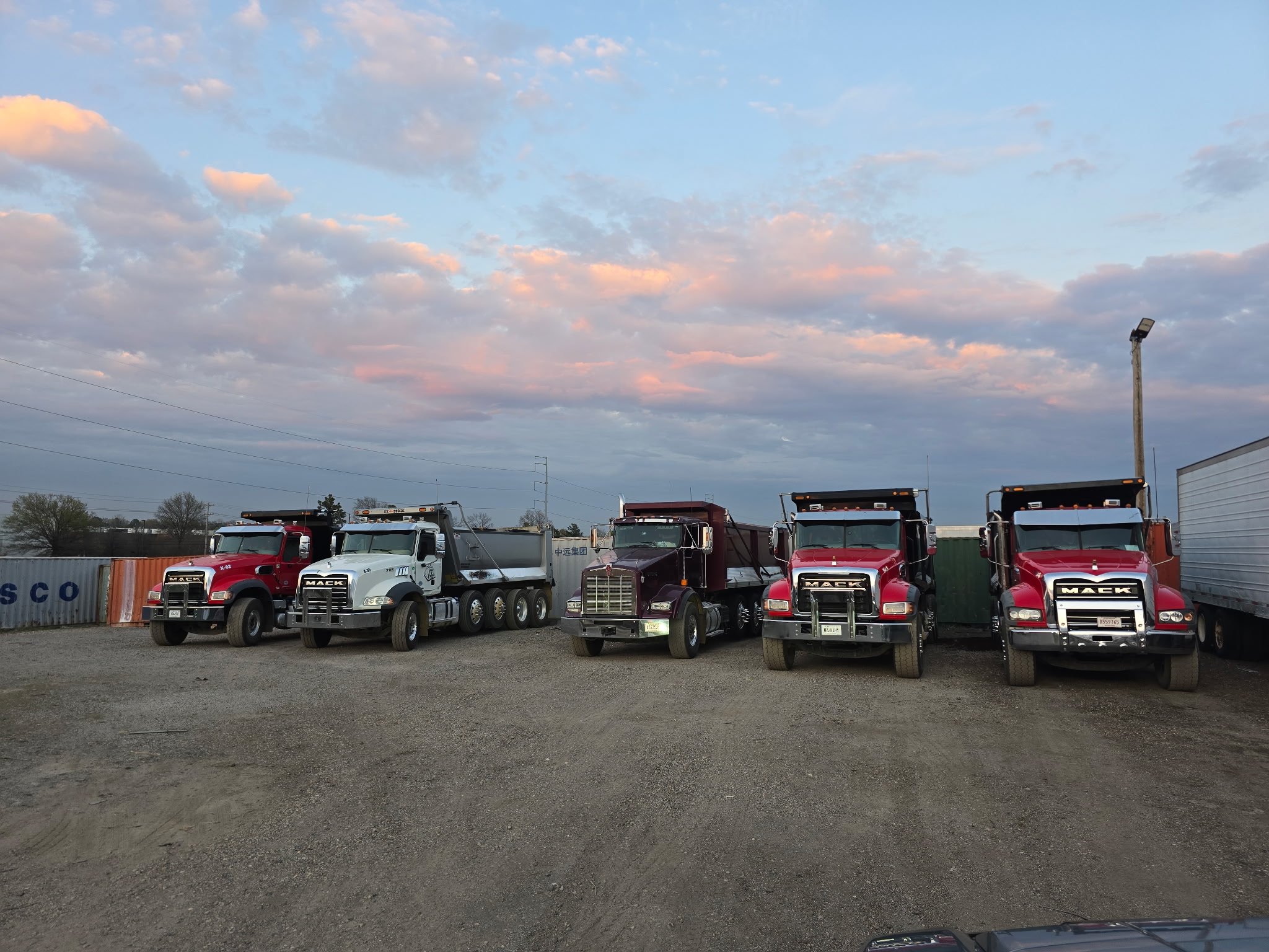 Multiple Tare 2 Tons Xpress dump trucks parked beneath a colorful evening sky
